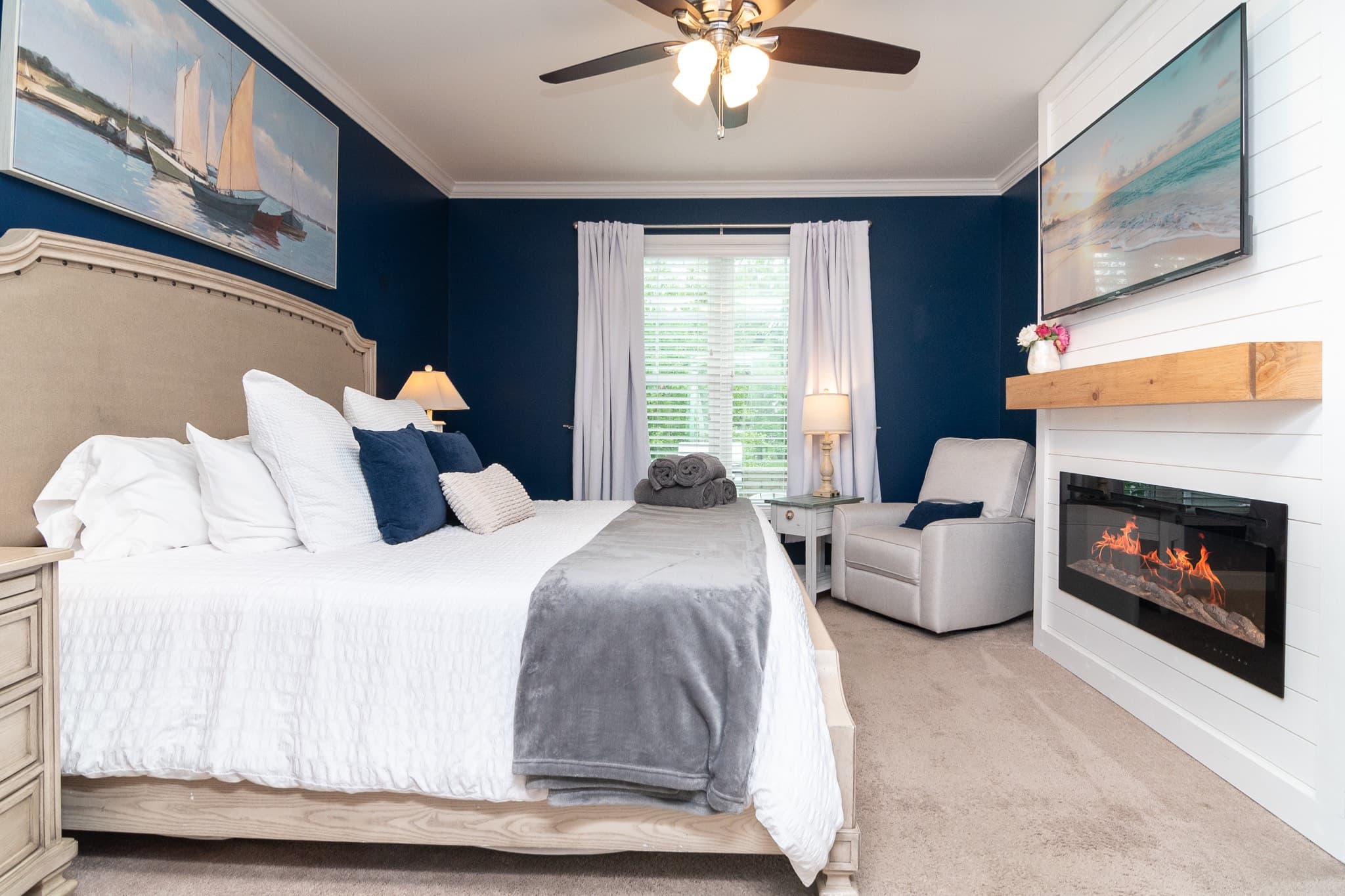 This first-floor master suite features a plush king bed, dresser, full-length mirror, recliner chair, electric fireplace, blackout curtains, a 55” Roku TV with cable, and a large closet. The room overlooks the private patio.